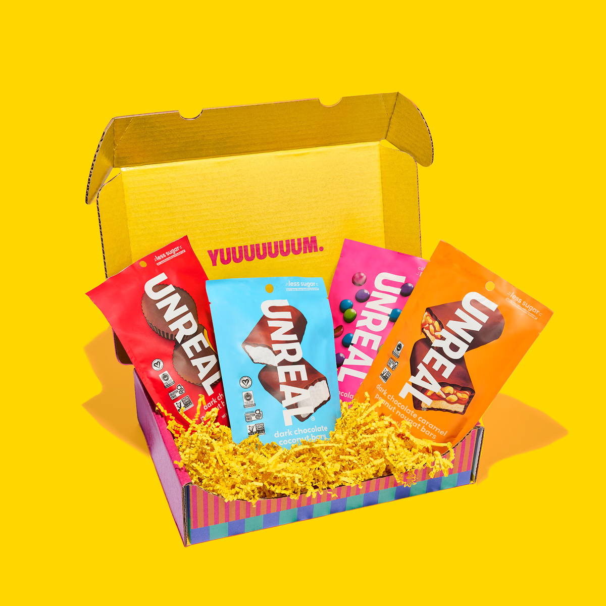 Angled top view of Unreal Gift Box with four UNREAL pouches on yellow crinkle paper and YUUUUUUM printed inside the lid