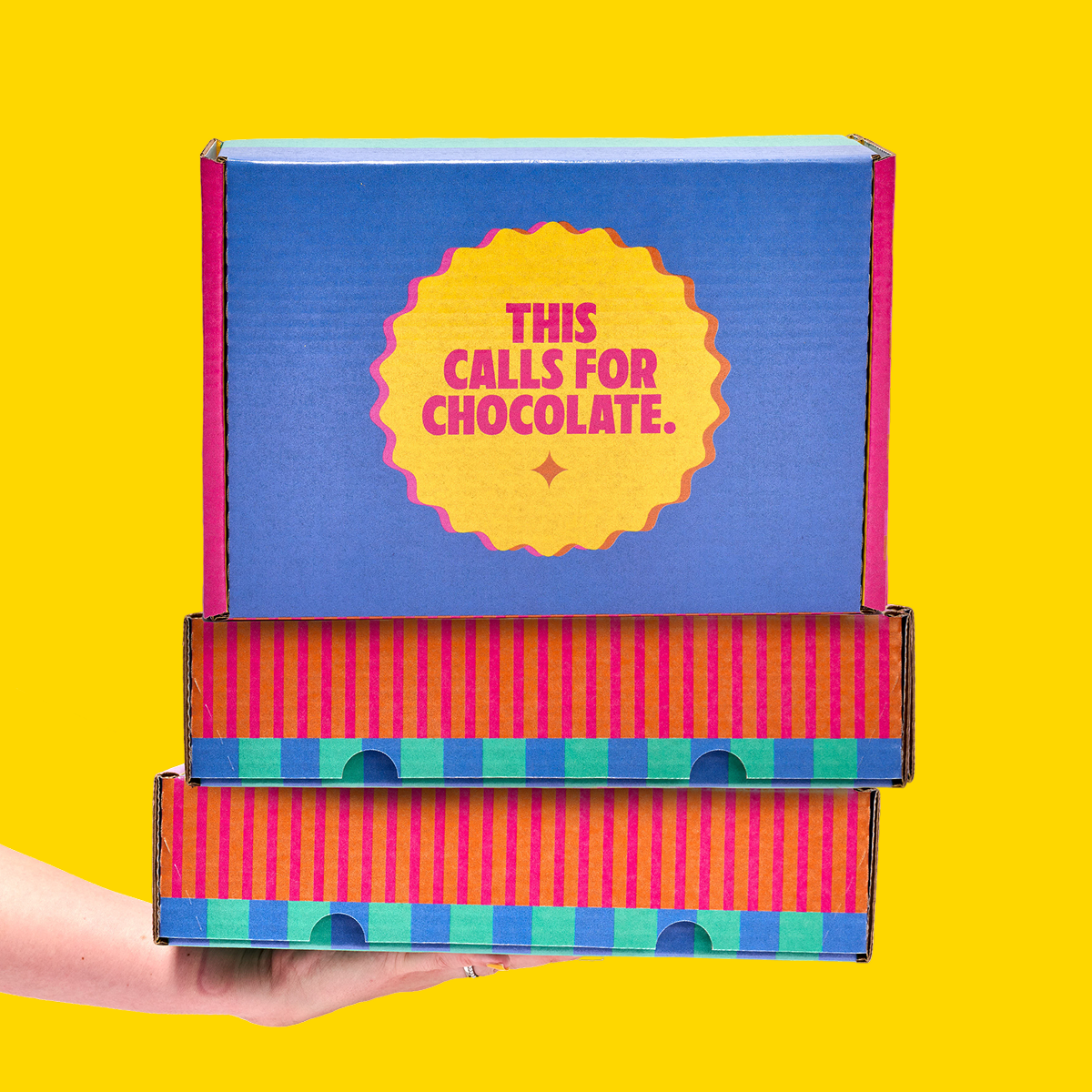 Front view of Unreal gift boxes with blue lid reading THIS CALLS FOR CHOCOLATE, held by hand against yellow background