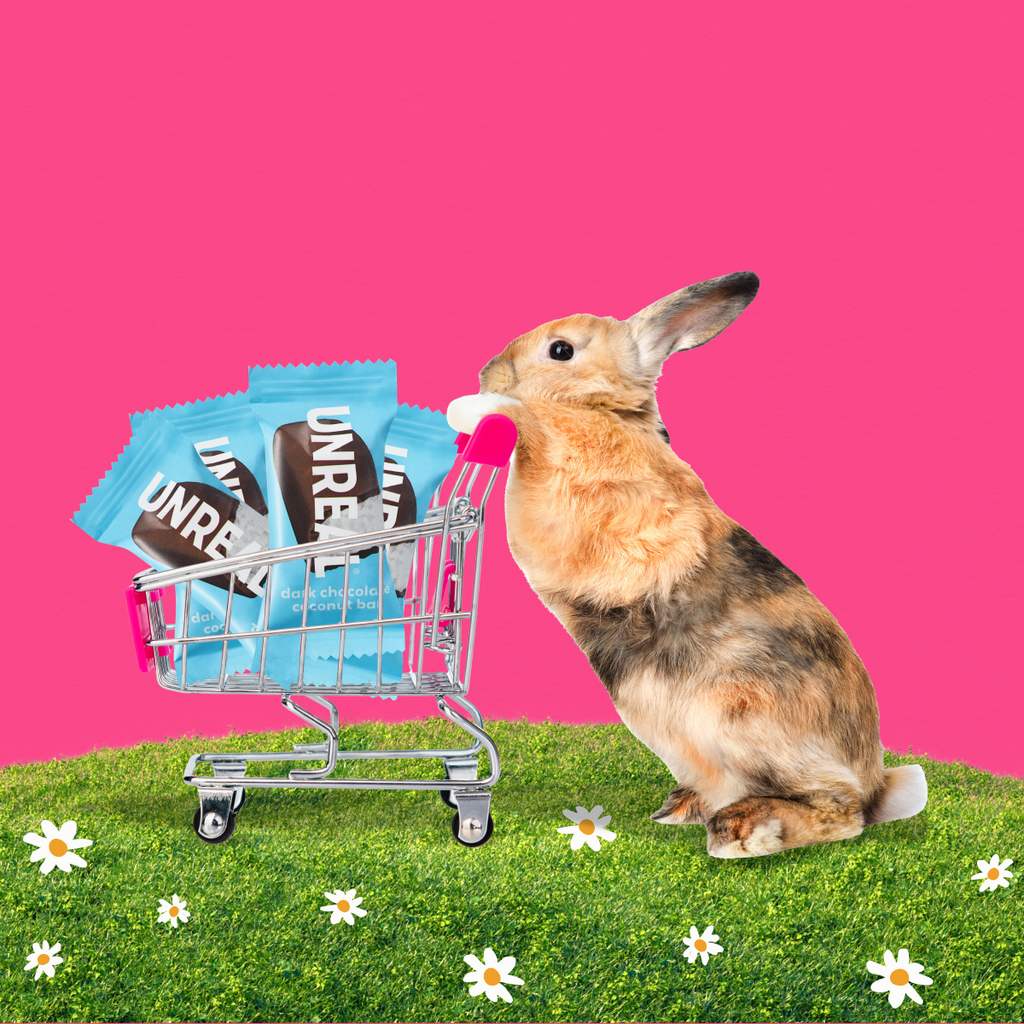 Brown-and-gray rabbit pushing a mini shopping cart of UNREAL dark chocolate coconut bar wrappers on grass with a pink background
