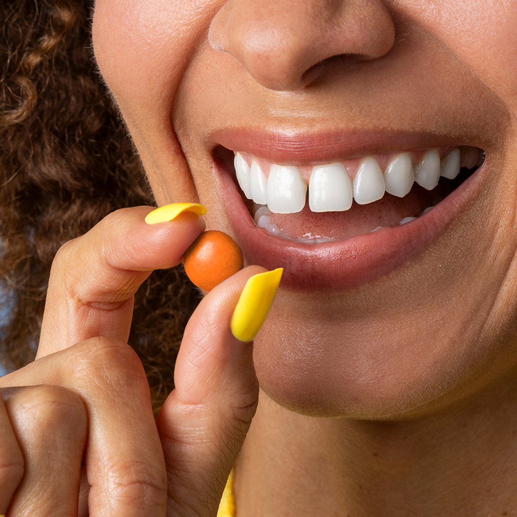 Close-up of Peanut Butter Drops held between fingers near a smiling mouth, showing orange candy shell for scale