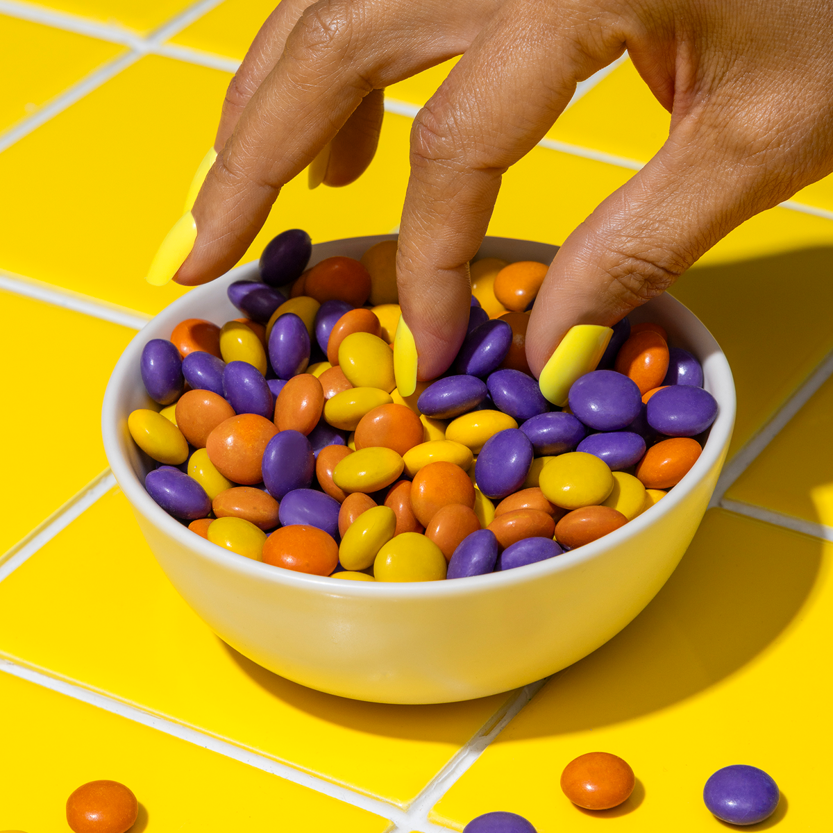 Close-up of Peanut Butter Drops in a white bowl, orange, purple, and yellow candy shells, hand with yellow nails picking one on yellow tiles