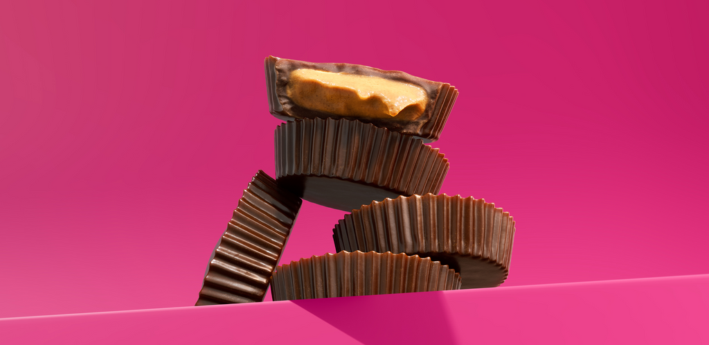 Stack of chocolate peanut-butter cups on bright pink background, one cut open showing creamy peanut butter filling.